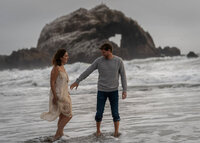 man reaching out to hold the woman's hand at sunset as they wade through the surf at the beach with a large rock in the background forming a shape of a heart