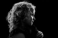 A black and white picture of a young blonde girl staring off, holding a stuffed animal. She is softly lit against a black background.