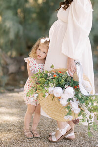 Young girl in a floral dress stands beside her pregnant mother during an outdoor family portrait session, holding a straw hat filled with fresh flowers in a soft, sunlit garden setting.