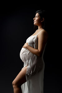 Artistic maternity portrait of a pregnant woman in a sheer white gown against a dark background photographed by Kaitlyn Dawn Photography.