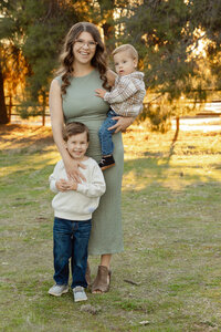 Mother standing in a sunlit field holding her baby while her young son stands beside her, candid lifestyle family portrait by Kaitlyn Dawn Photography, Yucaipa family photographer.