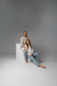 Parents sitting together on the studio floor in a relaxed pose against a gray backdrop, timeless indoor family portrait by Kaitlyn Dawn Photography.