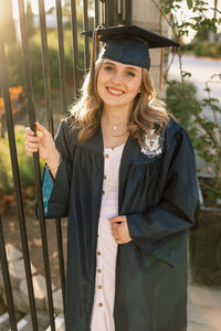 High school graduate in a cap and gown standing outdoors by an iron fence with natural golden hour lighting, graduation portrait by Kaitlyn Dawn Photography.
