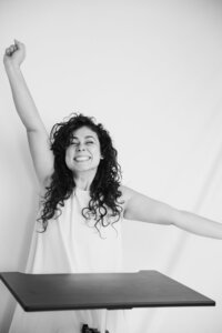 A black and white picture of a woman with dark curly hair smiling joyfully as she sits in front of a white backdrop. She wears a white dress and extends both arms away in a joyful stretch. A black table rests in front of her.