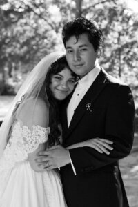 A bride and groom stand closely together, smiling softly. The bride wears a lace off-shoulder dress and veil, while the groom is in a dark suit with a boutonniere. They are outdoors with blurred greenery in the background. Black and white image.
