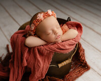Sleeping newborn baby girl wrapped in a rust-colored swaddle and posed in a basket prop during a studio newborn photography session with newborn photographer Kaitlyn Dawn Photography.