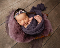 Sleeping newborn baby girl wrapped in a deep purple swaddle and posed in a basket with a soft mauve blanket during a professional studio newborn photography session by Yucaipa newborn photographer Kaitlyn Dawn Photography.