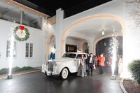 Guests alighting from a classic car at a brightly lit venue entrance decorated with a christmas wreath.