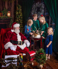 Children gathered around a glowing magical globe with Santa during a Santa’s Magical Christmas photography session in Jacksonville, Florida with Nature’s Child Photography, rich holiday decor and sparkling Christmas magic filling the cozy North Pole scene.