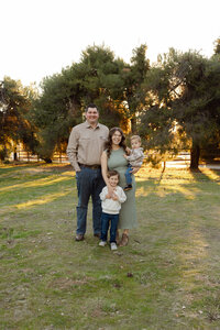 Family of four standing closely together outdoors in warm evening light, coordinated neutral outfits captured by Kaitlyn Dawn Photography, lifestyle outdoor family photographer in Yucaipa.