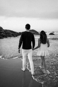 Black-and-white photo of a couple holding hands walking along the beach