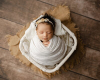 Sleeping newborn baby wrapped in a cream swaddle and posed in a rustic basket during a studio newborn photography session with newborn photographer Kaitlyn Dawn Photography.