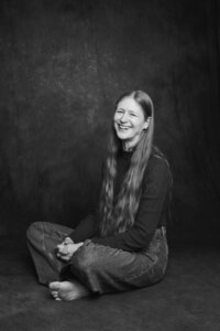 A black and white picture of a woman smiling, sitting cross legged on a dark gray backdrop.