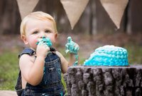 Blonde toddler in denim overalls with blue frosting on their hands and face during an outdoor cake smash in Mandarin. - A portrait experience by Nature's Child Photography.