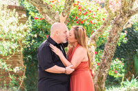 Couple kissing under a tree surrounded by red flowers during a romantic engagement photo session in a park near Pasadena.