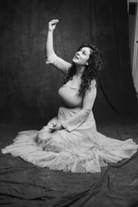 A black and white picture of a woman with dark curly hair sitting on a dark gray backdrop. She wears a long tulle skirt and lifts her gaze, speaking towards her raised hand.