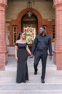 An elegant couple poses beneath the ornate red-brick arches of Flagler College in St. Augustine, Florida. She wears a floor-length off-the-shoulder black gown, and he’s dressed in an all-black outfit. Captured by Phavy Photography, a St. Augustine engagement photographer known for luxury, editorial-style sessions at historic and iconic locations.
