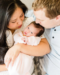Mother and father cuddling and kissing their newborn baby girl while she looks toward the camera during an in-home lifestyle session.