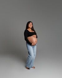 Pregnant woman standing alone in a black top and jeans, smiling while holding her baby bump during a maternity photoshoot in Yucaipa by Kaitlyn Dawn Photography.