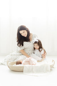 Mommy and me portrait of a mother in a Dallas photography studio sitting on the floor with her newborn baby in a basket while snuggled with her little girl by her as the family photographer captures them together.