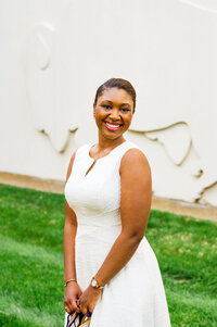 University graduate wearing white dress and smiling at the camera with the Belmont bison wall behind her