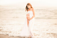 Family Photographer, a n expectant woman in her pregnancy holds her belly standing on the beach