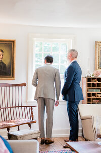 Grooms look out the sun lit window before the ceremony