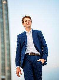 Senior portrait in downtown Mobile, Alabama— walking wear a blue suit with a skyscraper in the background.