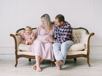 Mom, dad and toddler daughter sitting on a chaise during a maternity session in a Seattle studio
