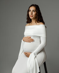 Pregnant mom seated in an elegant white dress, softly cradling her baby bump during a maternity photoshoot with Kaitlyn Dawn Photography in the Inland Empire.