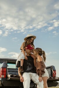 A portrait photo of a family taken from below showcases a blue sky with puffy clouds as they laugh, sitting on the back of their truck during family photos near Arvada, Colorado.