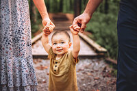 Baby holding both parents hands while walking along a wooded path during a lifestyle family session in North Carolina
