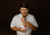 Father holding his newborn baby against a dark backdrop. Photographed by Inland Empire newborn photographer Kaitlyn Dawn Photography.