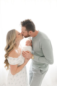 Mom and dad kissing , while holding their newborn together