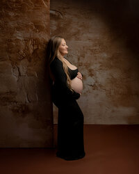 Expecting mother in a black maternity dress standing against a textured wall during a maternity portrait session by Kaitlyn Dawn Photography.