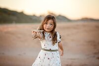 Young girl with windblown hair wearing a white floral dress holding out a shell toward the camera during a beach child portrait session. - A portrait experience by Nature's Child Photography.