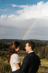 A newlywed couple standing close looking at each other with a rainbow in the distance.