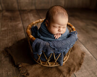 Sleeping newborn baby boy wrapped in a deep blue swaddle and posed in a rustic wicker basket on a wooden backdrop during a newborn photography session by Kaitlyn Dawn Photography.