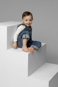 Smiling toddler girl sitting on modern white studio steps in a neutral gray setting, classic child portrait by Kaitlyn Dawn Photography, Yucaipa family photography studio.