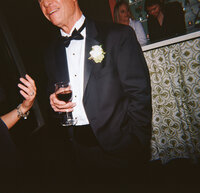 Close-up of a wedding guest in a black tuxedo holding a glass of red wine, with a white boutonniere pinned to his lapel, during the reception at Burritt on the Mountain.