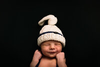 A naked baby with a crochet beanie sleeps against a black backdrop.