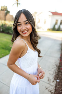 Smiling high school senior girl posing outdoors in a white dress with soft sunset lighting, senior portrait by Kaitlyn Dawn Photography.