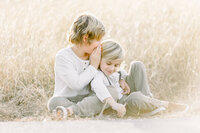 Two brothers sitting in a grassy Dallas field as the big brother whispers a secret to his little brother.