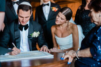 A groom, wearing a tuxedo, signs the ketubah at a Jewish wedding ceremony while the bride in a white gown watches