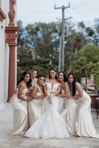 The bride and her bridesmaids pose gracefully outside the historic Lightner Museum in St. Augustine, Florida. The bride stuns in a form-fitting, off-the-shoulder lace gown with a dramatic train, while her bridesmaids wear matching champagne satin dresses that exude elegance and cohesion. The soft tones, architectural backdrop, and poised styling create a luxurious and elevated bridal portrait. Captured by Phavy Photography in a true-to-color, editorial documentary style that showcases refinement, beauty, and timeless sophistication.