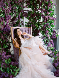 bride relaxing in a chair with flowing blush gown surrounded by a large lilac floral installation