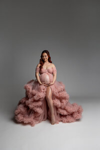 Elegant maternity portrait of a pregnant woman in a blush pink couture maternity gown holding her baby bump during a studio session by Kaitlyn Dawn Photography.