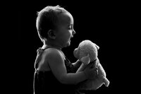 A black and white photograph of a baby laughing while holding a stuffed lamb. The lighting is dramatic, highlighting the edges of the baby and stuffed animal.