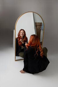 Red-haired woman smiling with makeup brush while seated at a desk. Photograph by Yucaipa branding photographer Kaitlyn Dawn Photography.