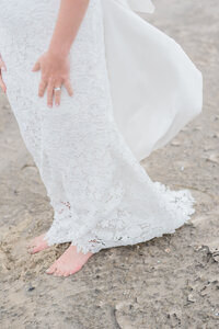 A close-up of the bride’s white lace wedding dress as she stands barefoot on the sandy beach.
